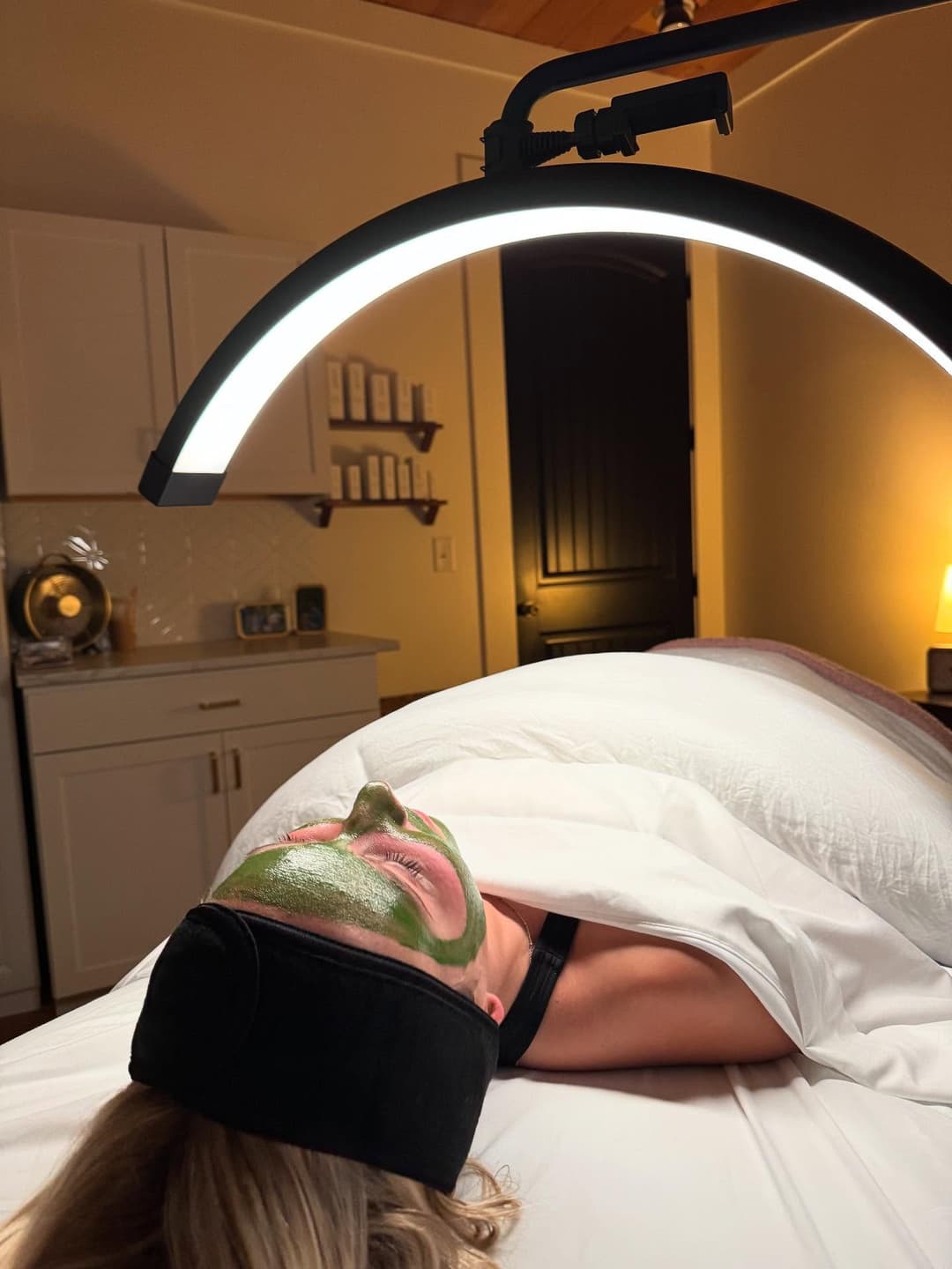 Woman receiving facial treatment with green mask under bright light in spa environment.