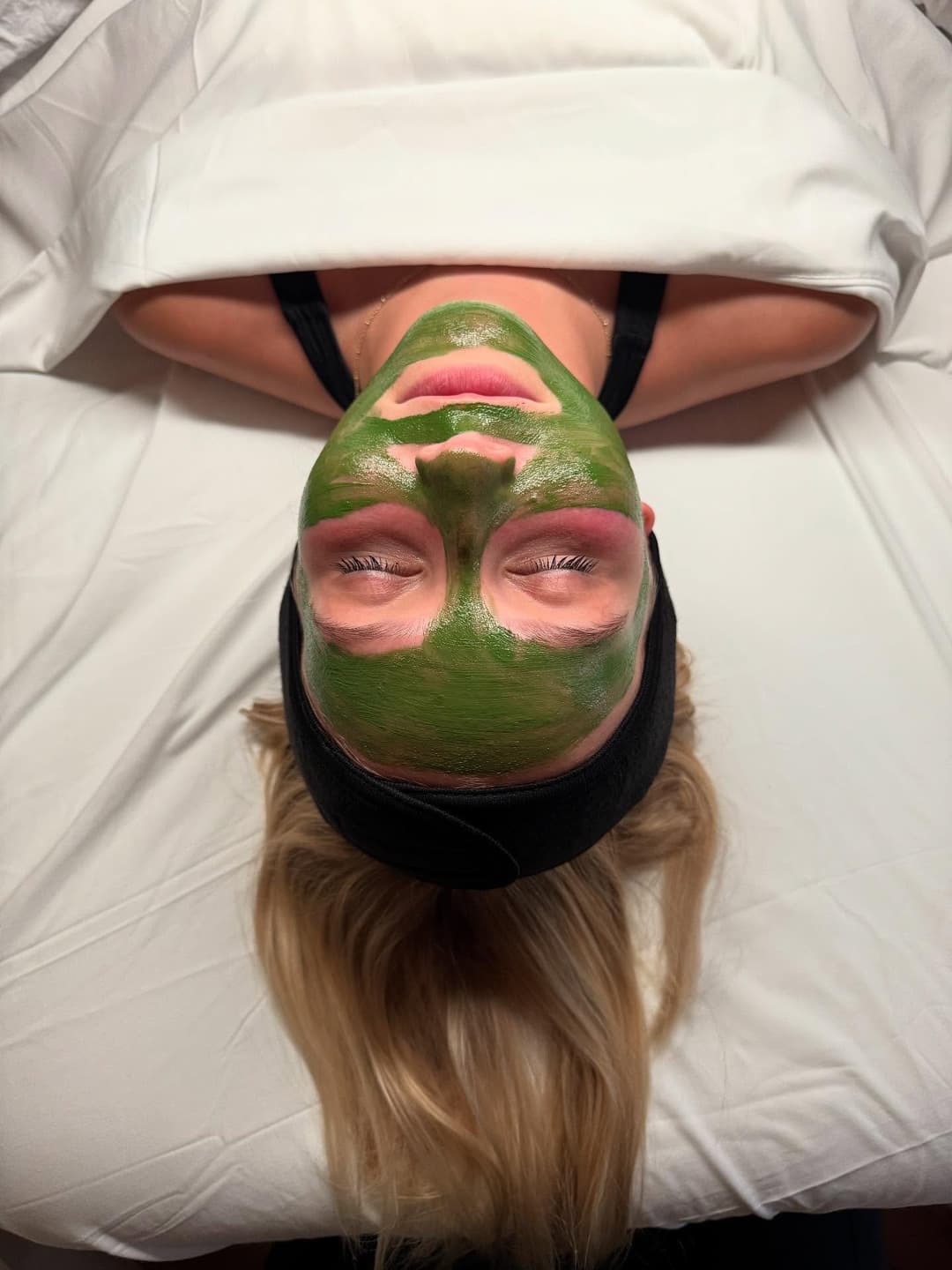 Woman receiving a green facial mask treatment in a spa setting, relaxing on a white table.