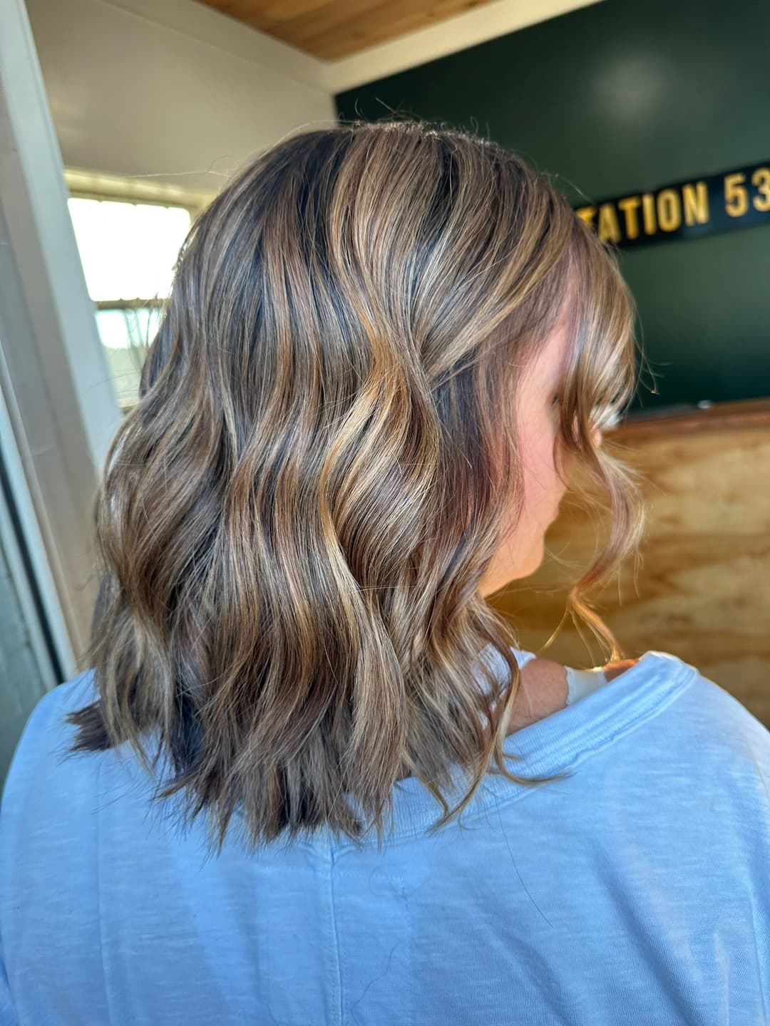 Woman with wavy, highlighted hair seen from behind in a stylish salon setting.