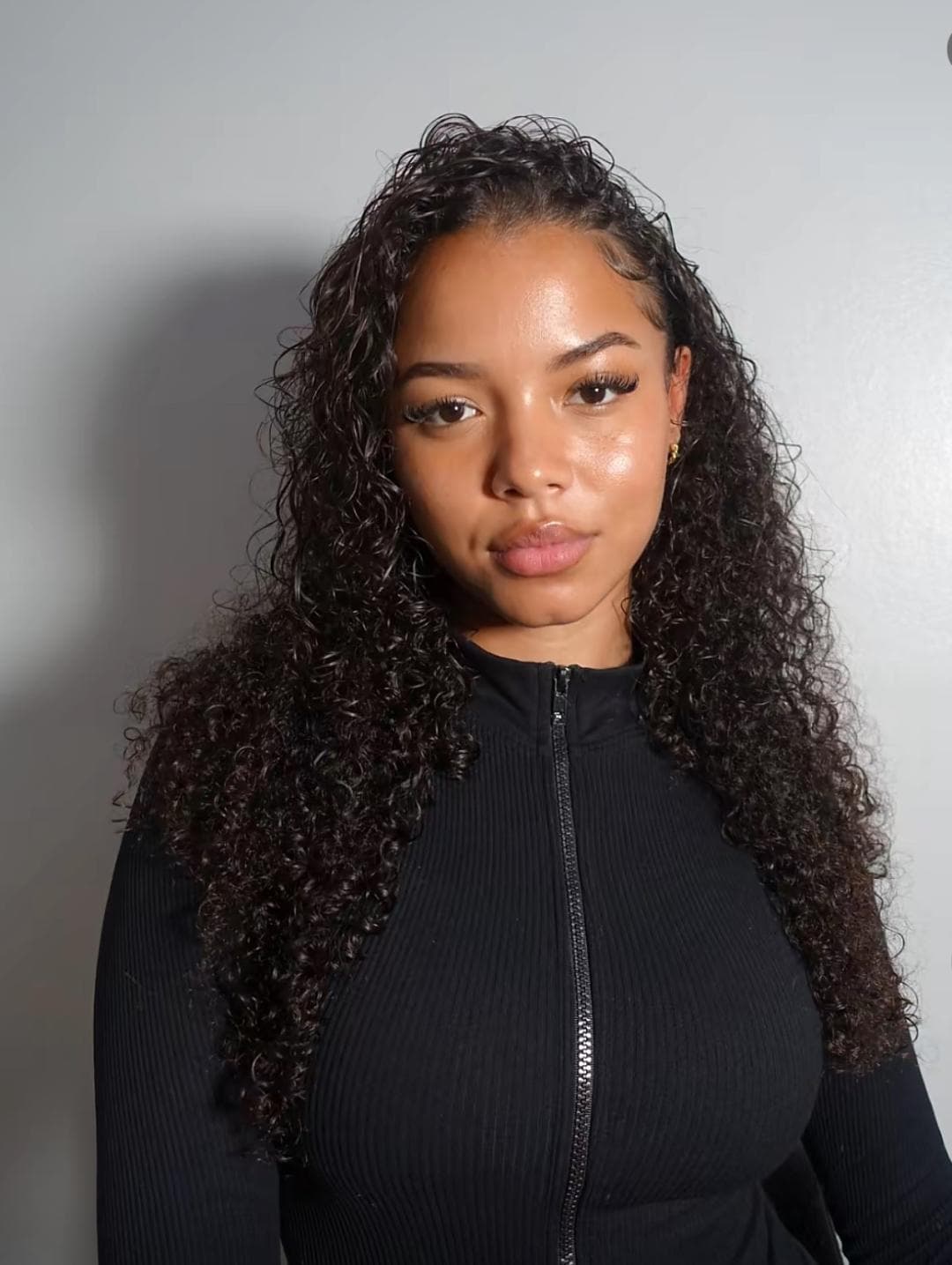 Woman with curly hair wearing a black zip-up top, posing against a light background.