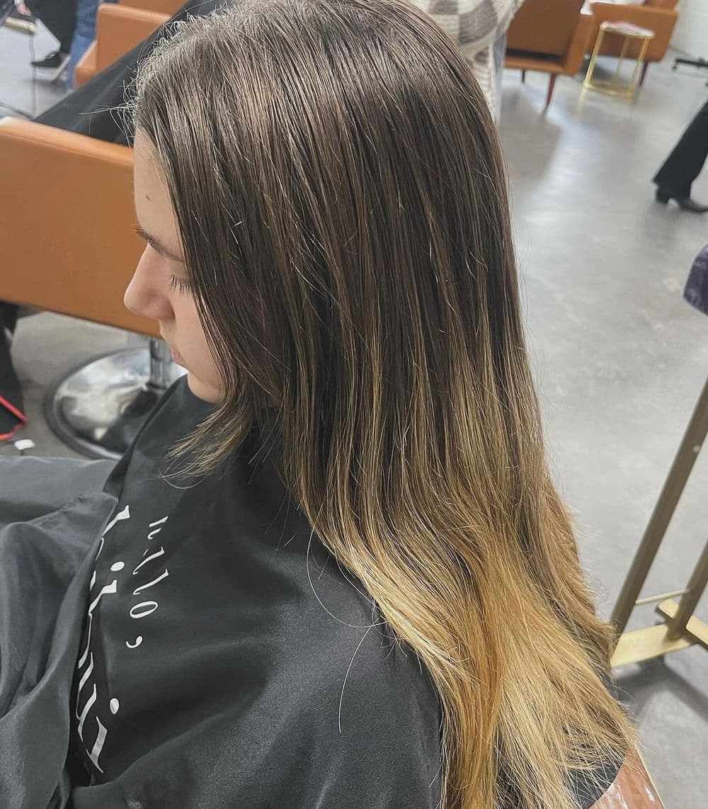 Woman with long hair transitioning from dark roots to lighter ends, seated in a salon.